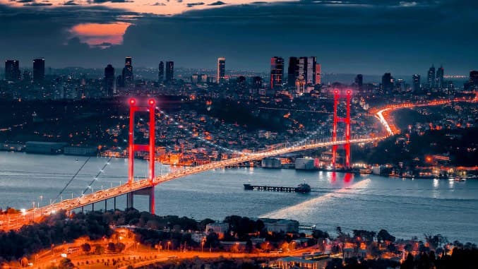 Alacakaranlıkta ışıklandırılmış Boğaziçi Köprüsü ve arkasında İstanbul silueti